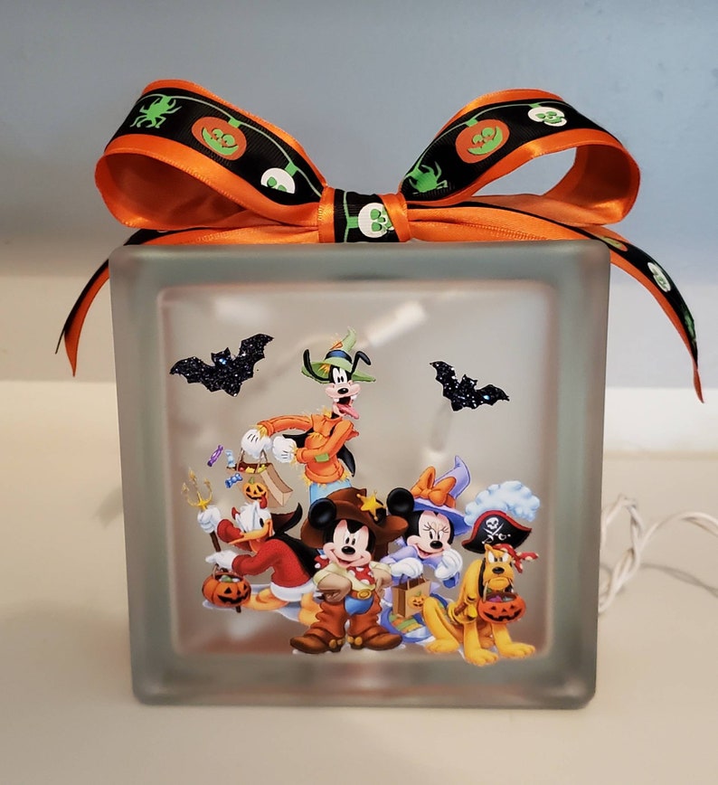 May include: A clear glass block with a Halloween-themed image of Mickey Mouse, Minnie Mouse, Goofy, Donald Duck, and Pluto dressed in costumes. The block is lit from within and has a black and orange ribbon tied around the top.