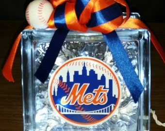 Puede incluir: Un bloque de vidrio con un logotipo de los New York Mets y una pelota de béisbol en la parte superior. El bloque está decorado con una cinta naranja y azul.