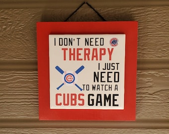 May include: A red wooden sign with a white background and the text "I don't need therapy I just need to watch a Cubs game". The sign has a baseball bat and ball graphic with the Cubs logo.
