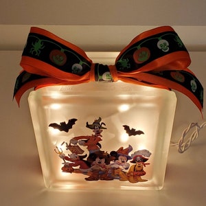 May include: A light-up glass block with a Halloween-themed image of Mickey Mouse and friends dressed in costumes. The block is decorated with an orange and black ribbon with a skull and pumpkin design.