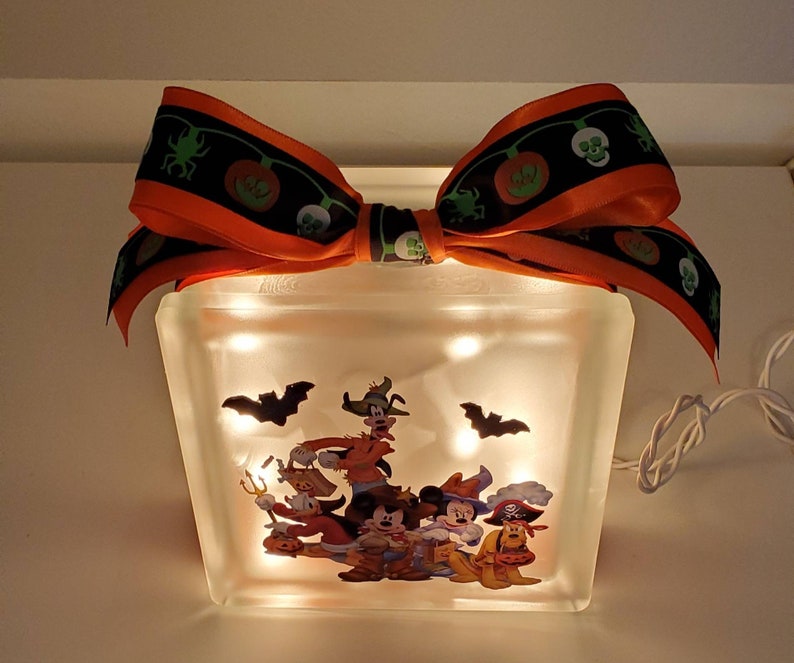 May include: A light-up glass block with a Halloween-themed image of Mickey Mouse and friends dressed in costumes. The block is decorated with an orange and black ribbon with a skull and pumpkin design.