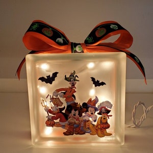 May include: A light-up glass block with a Halloween-themed image of Mickey Mouse and friends dressed in costumes. The block is decorated with a large orange bow with black and green ribbon.