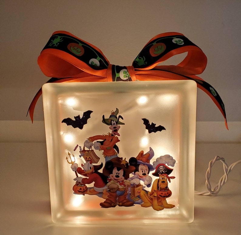 May include: A light-up glass block with a Halloween-themed image of Mickey Mouse and friends dressed in costumes. The block is decorated with a large orange bow with black and green ribbon.