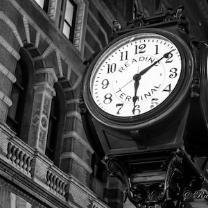 May include: Black and white photo of a clock face with the words "Reading Terminal" on it. The clock is mounted on a building with a brick facade.
