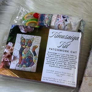 May include: A Kinusaiga Kit for a patchwork cat, packaged in clear plastic. The kit includes a foam board, scissors, a seam ripper, and various fabric strips. The label features a cat illustration and text describing the no-sew fabric art technique.