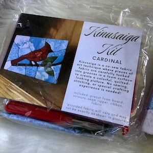 May include: A Kinusaiga Kit Cardinal craft set in a clear plastic bag. The kit includes a foam board, fabric pieces, scissors, a seam ripper, and ribbon. The packaging features a picture of a red cardinal bird on a branch and descriptive text.