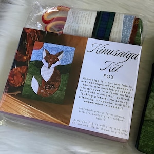 May include: A Kinusaiga Kit for creating a fox image, featuring a rectangular box with a fox illustration, text describing the no-sew fabric technique, and a clear plastic bag containing scissors, a seam ripper, ribbon, and various fabric pieces.