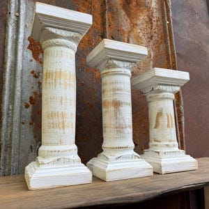 May include: Set of three decorative wooden pillars with a distressed white finish. The pillars have a classic column design with a square top and base, and varying heights. The wood grain is visible, adding to the rustic charm.