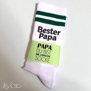 May include: White socks with green stripes near the top, featuring the text "Bester Papa" and "Papa, Du bist die coolste Socke" in black. A light green band wraps around the sock.