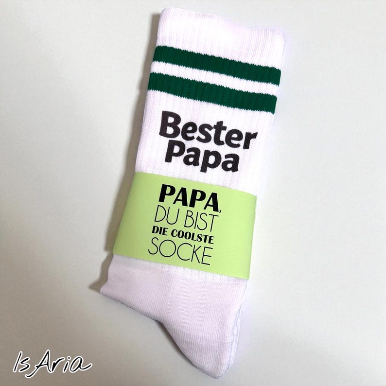 May include: White socks with green stripes near the top, featuring the text "Bester Papa" and "Papa, Du bist die coolste Socke" in black. A light green band wraps around the sock.