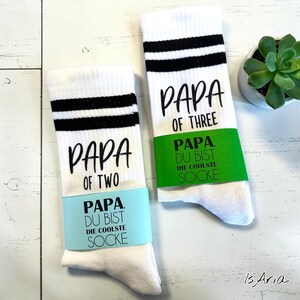 Könnte beinhalten: Zwei Paar weiße Socken mit schwarzen Streifen oben. Ein Paar sagt "PAPA OF TWO" und das andere "PAPA OF THREE". Jedes Paar hat ein Band mit "PAPA DU BIST DIE COOLSTE SOCKE".