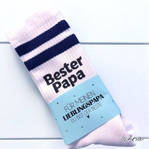 May include: White athletic socks with navy blue stripes near the top. The words "Bester Papa" are printed in black. A light blue band wrapped around the socks reads "F&Uuml;R MEINEN LIEBLINGSPAPA DU BIST DER BESTE".
