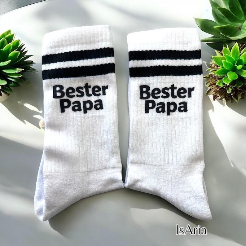 May include: A pair of white socks with black stripes near the top. The text "Bester Papa" is printed in black on each sock. The socks are displayed on a white surface.