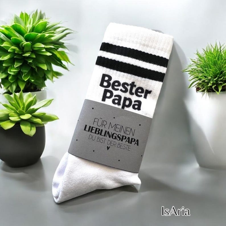 May include: White socks with black stripes and the words "Bester Papa" printed in bold black letters. A gray label reads "F&Uuml;R MEINEN LIEBLINGSPAPA DU BIST DER BESTE". The socks are folded and presented with small potted plants in the background.
