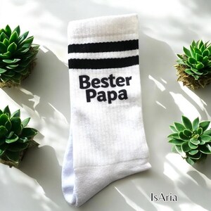 May include: White socks with black stripes and the text "Bester Papa" in black. The socks are surrounded by green succulents.