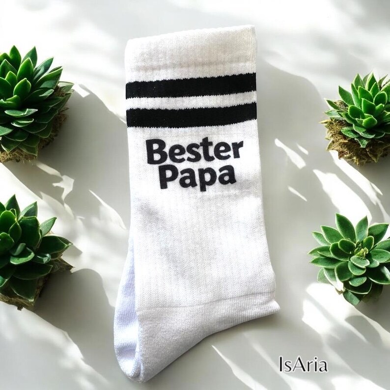 May include: White socks with black stripes and the text "Bester Papa" in black. The socks are surrounded by green succulents.