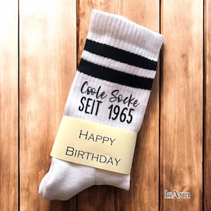 Könnte beinhalten: Weiße Socken mit schwarzen Streifen und dem Text "Coole Socke SEIT 1965". Ein gelbes Band um die Socken zeigt "HAPPY BIRTHDAY". Die Socken liegen auf einer Holzoberfläche.