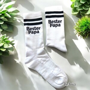 May include: A pair of white socks with black stripes at the top and the words "Bester Papa" printed in black. The socks are displayed on a white surface with green plants in the background.