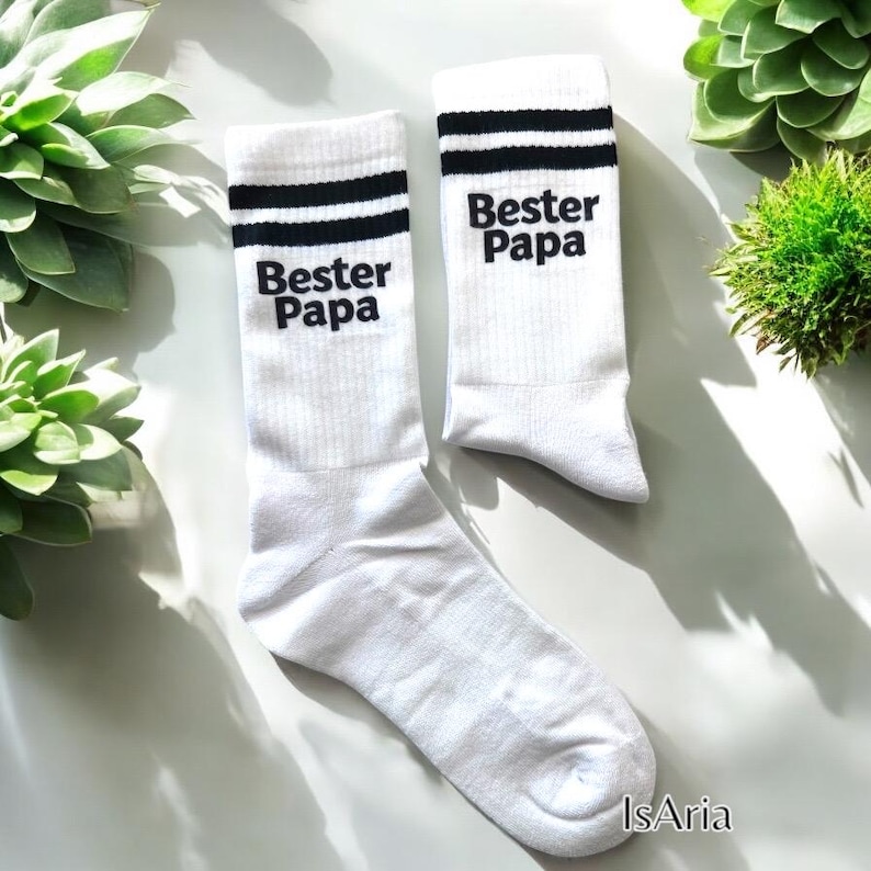 May include: A pair of white socks with black stripes at the top and the words "Bester Papa" printed in black. The socks are displayed on a white surface with green plants in the background.