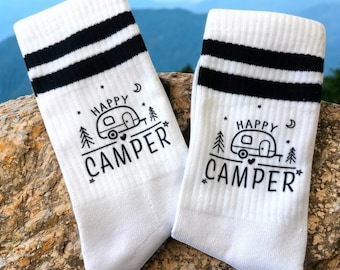Puede incluir: Un par de calcetines blancos con rayas negras en la parte superior. Los calcetines presentan un gráfico de una caravana con las palabras "HAPPY CAMPER" en negro. Los calcetines se muestran sobre una roca, con montañas al fondo.