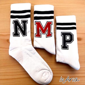 Könnte beinhalten: Drei weiße Socken mit schwarzen Streifen oben und großen Blockbuchstaben. Die Buchstaben sind schwarz, rot und schwarz und buchstabieren die Buchstaben N, M und P. Die Socken liegen auf einer Holzoberfläche.