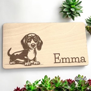 Könnte beinhalten: Ein Holzschild mit einer Dackel-Silhouette und dem Namen "Emma" eingraviert. Das Schild ist hellbraun und hat eine natürliche Holzmaserung.