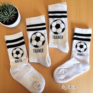 Könnte beinhalten: Vier Paar weiße Sportsocken mit schwarzen Streifen und Fußballgrafiken. Zwei Paare tragen die Aufschrift "TRAINER", eines "MATTIS" und eines "HANNES". Eine kleine Topfpflanze befindet sich oben links. Die Socken liegen auf einer hellbraunen Oberfläche.