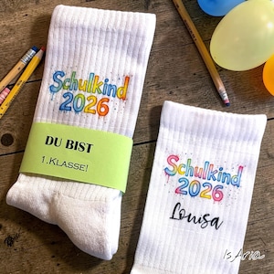 Könnte beinhalten: Zwei weiße Socken mit dem Text "Schulkind 2026" in bunten Buchstaben. Eine Socke hat den Text "DU BIST 1.KLASSE!" auf einem grünen Band. Die andere Socke hat den Namen "Louisa" darunter.