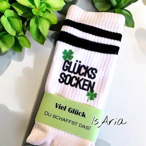Könnte beinhalten: Weiße Socken mit schwarzen Streifen und dem Schriftzug "GLÜCKS SOCKEN" in Schwarz, sowie grüne Kleeblatt-Grafiken. Ein grünes Band um die Socken trägt die Aufschrift "Viel Glück Du SCHAFFST DAS!"