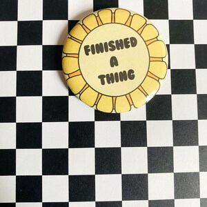 May include: A round, yellow button with a scalloped edge. The button features the text "FINISHED A THING" in a bold, black font. The background is a black and white checkered pattern.