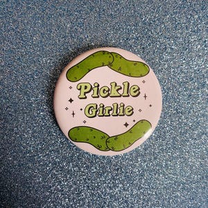 May include: A round, light pink button with the words "Pickle Girlie" in green, surrounded by two green pickle illustrations and four small black star shapes. The button is set against a sparkly, light blue background.