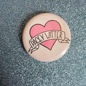 Könnte beinhalten: Ein runder Button mit einem rosa Herz und einem Banner mit der Aufschrift "PACEY WITTER" in schwarzer Schrift. Der Button hat einen weißen Hintergrund und liegt auf einer glitzernden blauen Oberfläche.