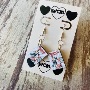 Könnte beinhalten: Ohrringe zum Aufhängen mit einem kleinen rechteckigen Bild eines Mannes auf einem roten Fahrrad. Die Ohrringe sind an einer weißen Karte mit schwarzen Herzen und dem Text "WCM" befestigt. Die Ohrringe haben silberfarbene Haken.