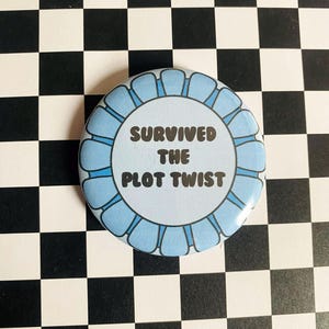 May include: A round, light blue button with the text "SURVIVED THE PLOT TWIST" in a bold, black font. The button has a scalloped edge and is set against a black and white checkered background.