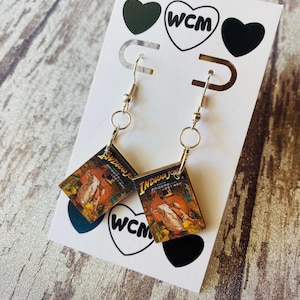 May include: Silver-toned dangle earrings featuring miniature book covers with the "Indiana Jones" movie poster design. The earrings are attached to a white card with black heart and "WCM" logo details.