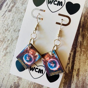 May include: A pair of dangle earrings featuring a rectangular image from the movie Labyrinth. The earrings have a silver-colored hook and chain design. The image shows a person holding a crystal ball. The earrings are attached to a white card with black heart and WCM logos.