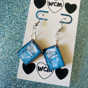 以下が含まれることがあります： シルバーカラーのドロップピアス。各イヤリングには、映画のキャラクターの青と白の画像が入った小さな正方形のペンダントが付いています。イヤリングは、黒いハートと「WCM」のデザインが施された白いカードに表示されています。