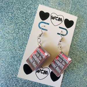 May include: Silver-tone earrings with miniature book charms, each showing the cover of "Interview with the Vampire" by Anne Rice. The earrings are suspended from silver-tone ear wires and presented on a white card with heart and logo details.