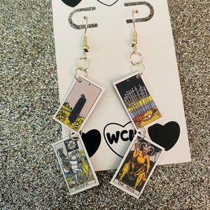 May include: Gold-coloured dangle earrings with tarot card designs. The cards show images of Death, The Devil, and other tarot symbols. The earrings hang from gold-coloured hooks and are displayed on a white card with black heart and "WCM" logo.