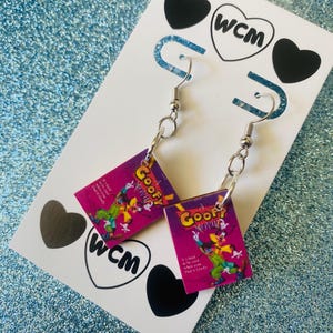 May include: A pair of dangle earrings featuring the cover art from the animated film "A Goofy Movie." The earrings have a purple background with the movie title in yellow and a cartoon image of Goofy. The earrings are attached to silver-colored fish hook ear wires.