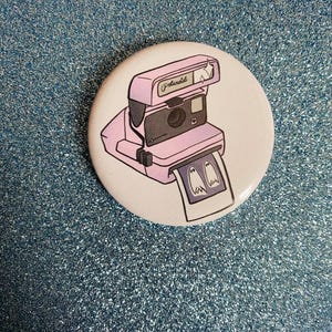 Könnte beinhalten: Ein runder Button mit einer rosa Polaroid-Kamera-Illustration. Die Kamera wirft ein Foto mit zwei Geisterfiguren aus. Das Wort "Polaroid" ist auf der Kamera aufgedruckt. Der Button befindet sich auf einer glitzernden blauen Oberfläche.