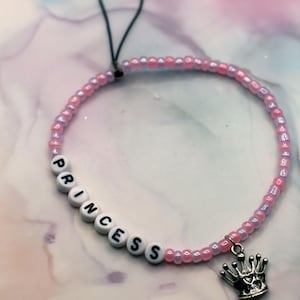 Puede incluir: Una pulsera de cuentas rosas y moradas. La palabra "PRINCESS" está escrita con cuentas de letras blancas. Un dije de corona plateada cuelga de la pulsera. La pulsera tiene un lazo negro adjunto.