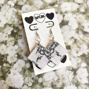 May include: A pair of square dangle earrings featuring the My Chemical Romance The Black Parade album cover art in black and white. The earrings are attached to a white display card with two black hearts and the text WCM inside a circle.