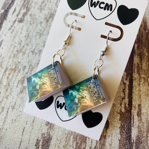 May include: A pair of dangle earrings featuring miniature book replicas. Each earring displays the cover of "Percy Jackson" with a teal and gold color scheme. The earrings hang from silver-colored fish hook ear wires and are attached to a white card with black heart and "wcm" logo.
