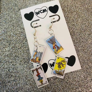 May include: Gold-tone earrings featuring four tarot card pendants. The cards depict various scenes with figures and symbols. The earrings hang from a white card with black heart and "WCM" logo.