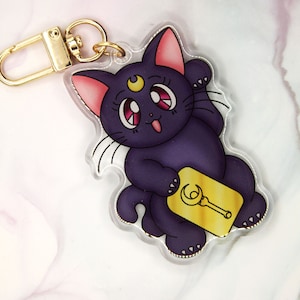 May include: A clear acrylic keychain featuring a cartoon black cat with pink ears, a crescent moon on its forehead, and a gold key. The keychain has a gold-colored clasp.