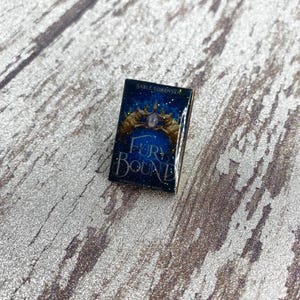 Puede incluir: Un pin esmaltado rectangular con un diseño de portada de libro sobre un fondo azul oscuro estrellado. El texto dice Sable Sorensen y Fury Bound. Dos figuras animales doradas flanquean una corona central con una gema morada brillante.