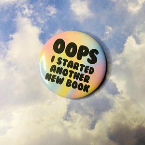 May include: A round button with a pastel rainbow background and the words "OOPS I STARTED ANOTHER NEW BOOK" in black lettering. The button is a fun accessory for book lovers.