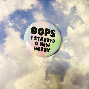 May include: A round button with the text "OOPS I STARTED A NEW HOBBY" in black font. The button has a pastel rainbow gradient background of pink, green, and yellow. The background is a cloudy sky.