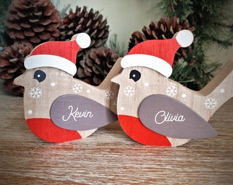 May include: Two wooden bird ornaments with red and white Santa hats, snowflake details, and gray wing accents. One bird has the name "Kevin" and the other "Olivia" in white script. Festive holiday decor.